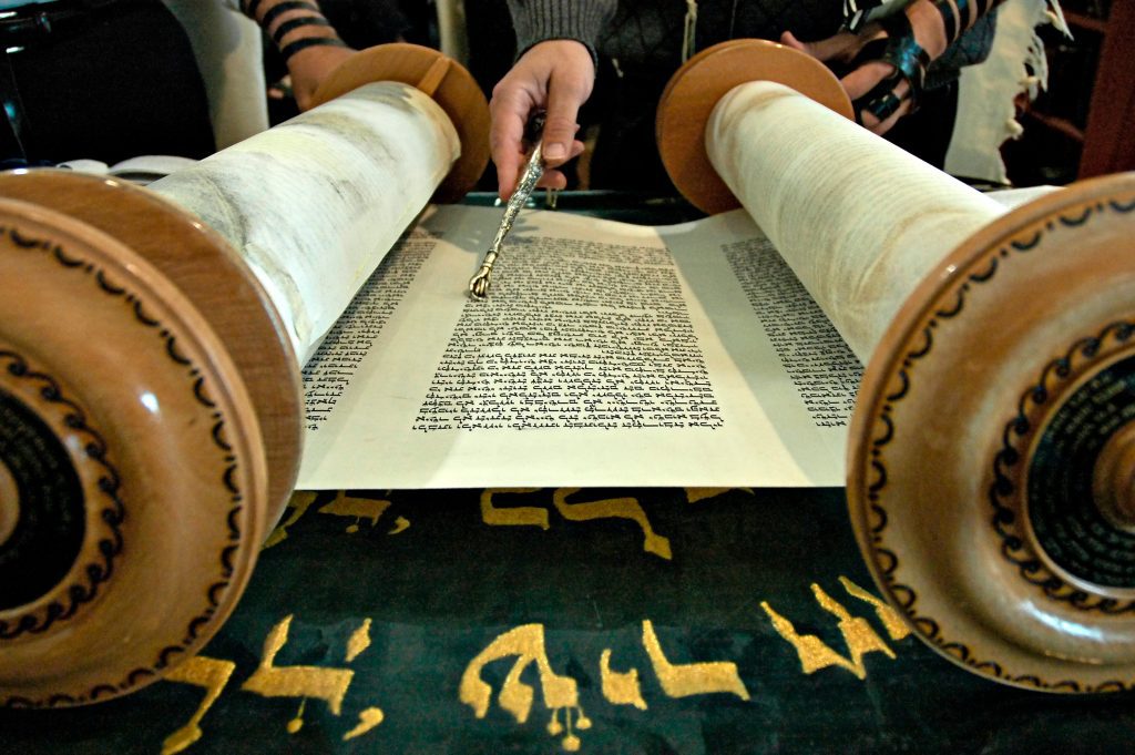 A Torah scroll opened in a synagogue with a hand holding a silver pointer.